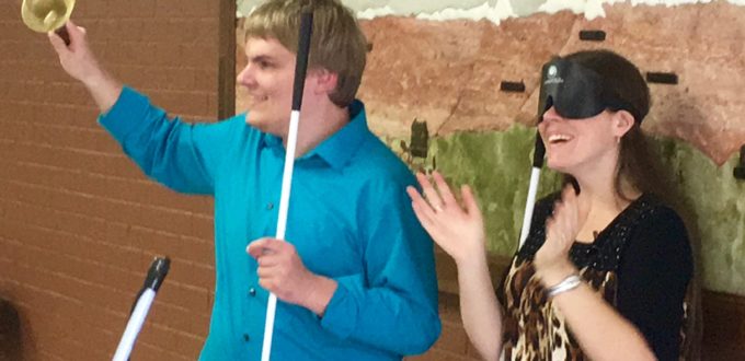 Man holding his bell high as he rings it, a woman stands beside him applauding