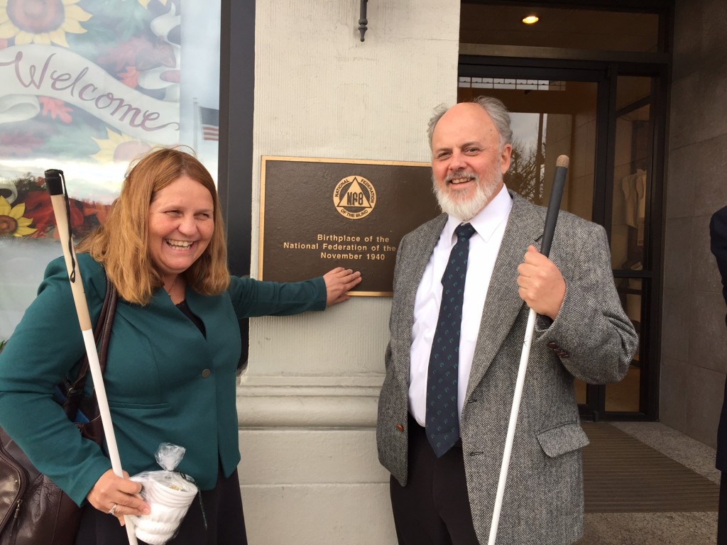 A smiling man and woman stand on each side of a bronze plaque with print and Braille visible. Both hold long white canes.