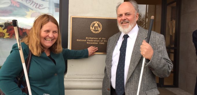 A smiling man and woman stand on each side of a bronze plaque with print and Braille visible. Both hold long white canes.