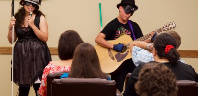 A young woman and young man with guitar perform before a seated crowd. They wear stunning hats.