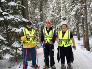Three people wearing snow shoes on a wintery forested path with snow covered branches.