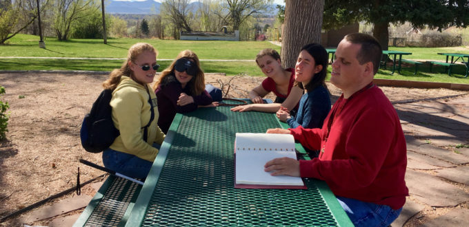 Five people around a picnic table, one reading Braille aloud to the others.