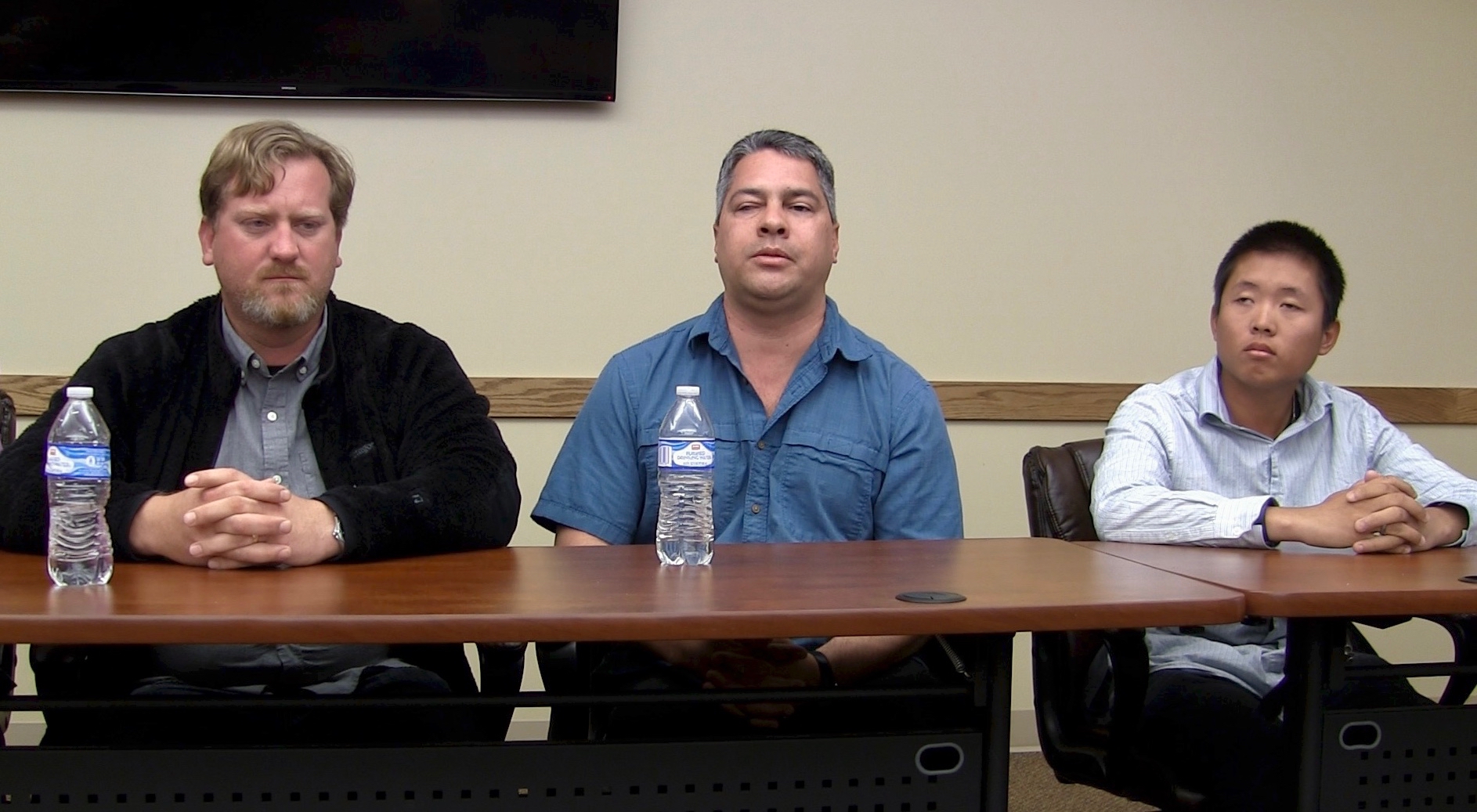 Three men seated at a long table, facing a room of listeners.