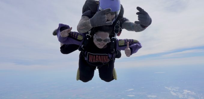 a grinning young woman strapped to her jump-master gives a double thumbs-up