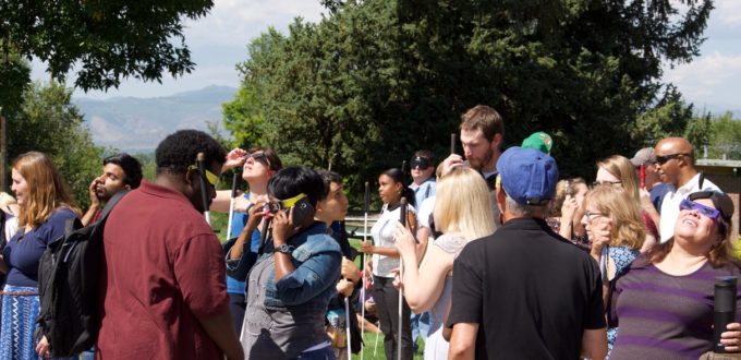 A Large group of CCB staff and students outside observing the eclipse. Some are wearing eclipse glasses while they listen to a live audio description
