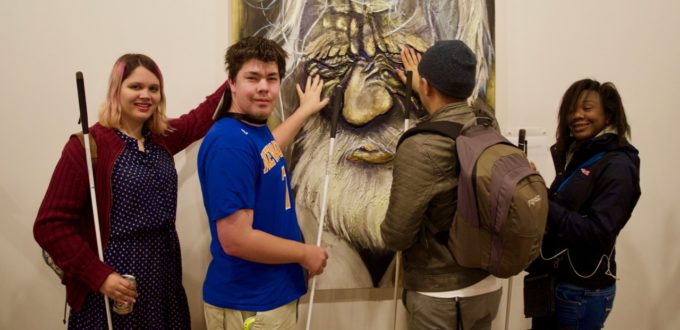 Adia, Mason M. and Cezar look at the Old Man Wincing while Ravi reads the Braille description