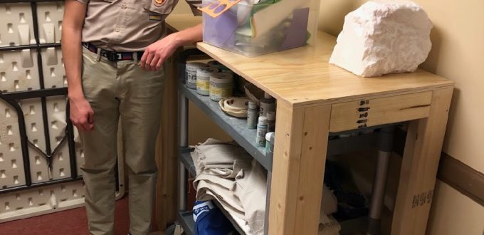 Alex LaBarre stands next one of the newly completed work benches showing how a rolling art supply cart fits underneath