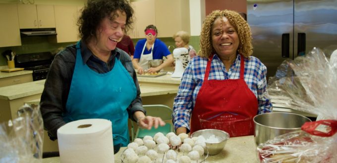 Blanca and Priscilla wearing festive colored aprons prepare a rack of home made powdered sugar cookies