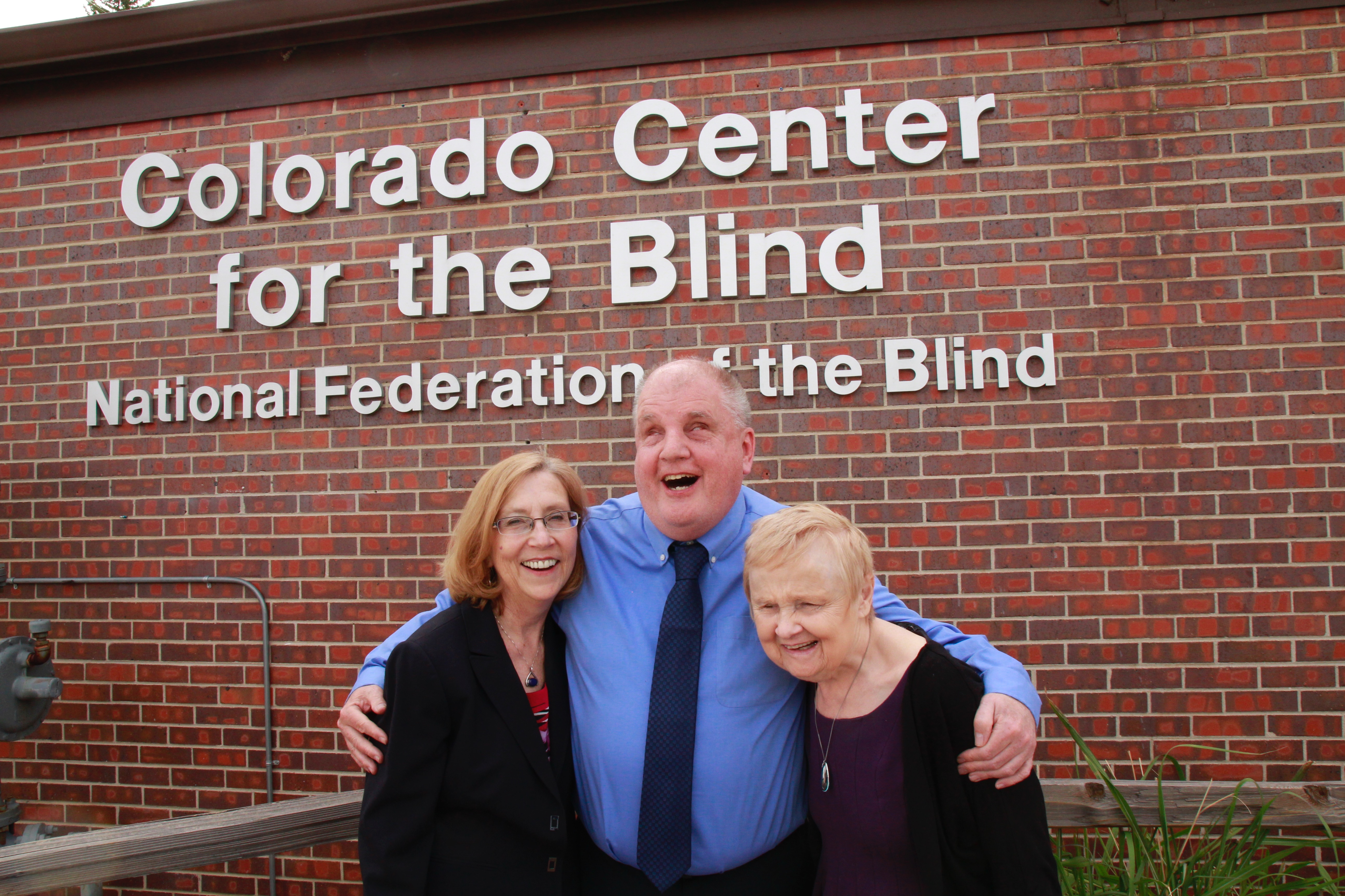 Duncan, Tom and Diane hug in front of the CCB sign on a bright spring day.