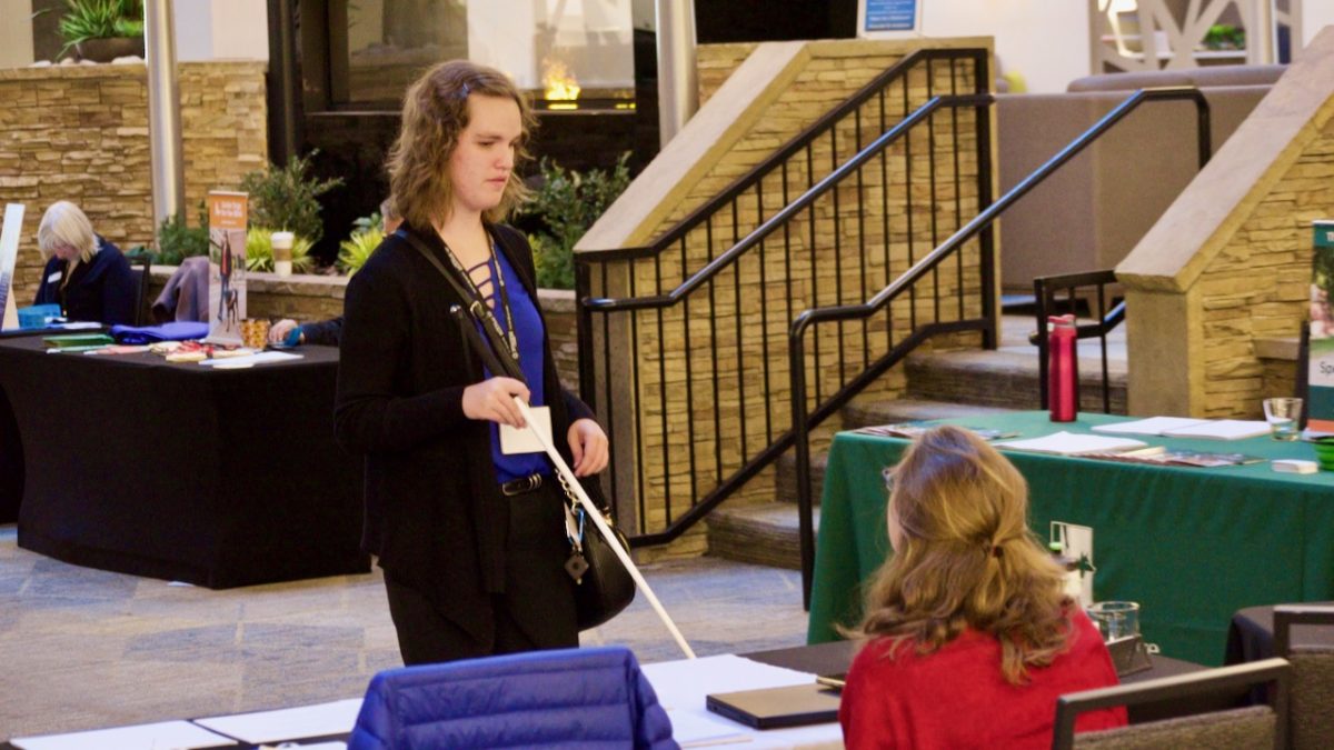 A young woman holding a white cane faces us as she talks to a vendor.