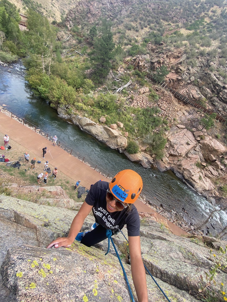 Carolina seen from above, smiling as she reaches the top of the rock, a wide view of fellow students below and across Clear Creek Canyon.